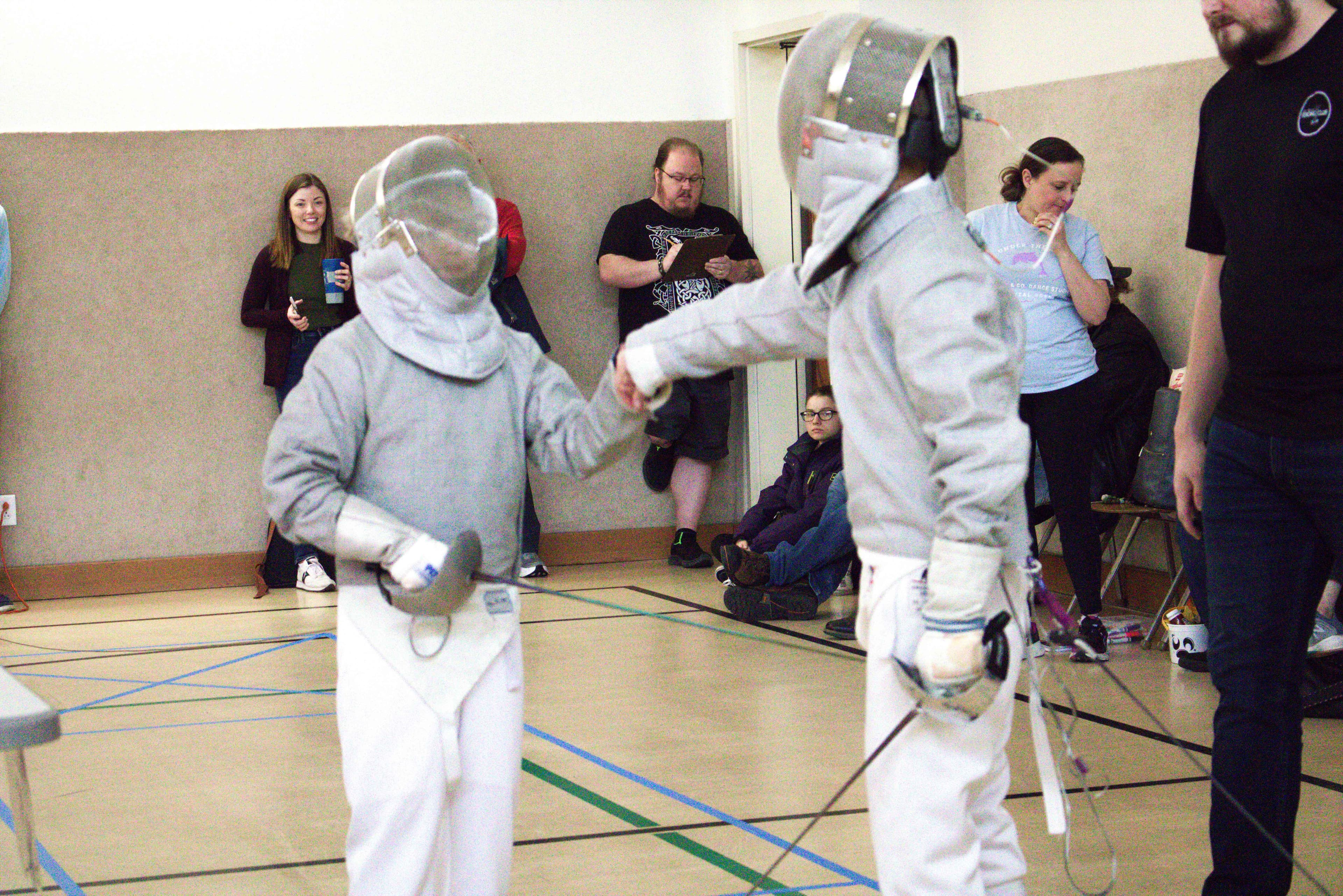 Two young club fencers facing off in their whites as members look on