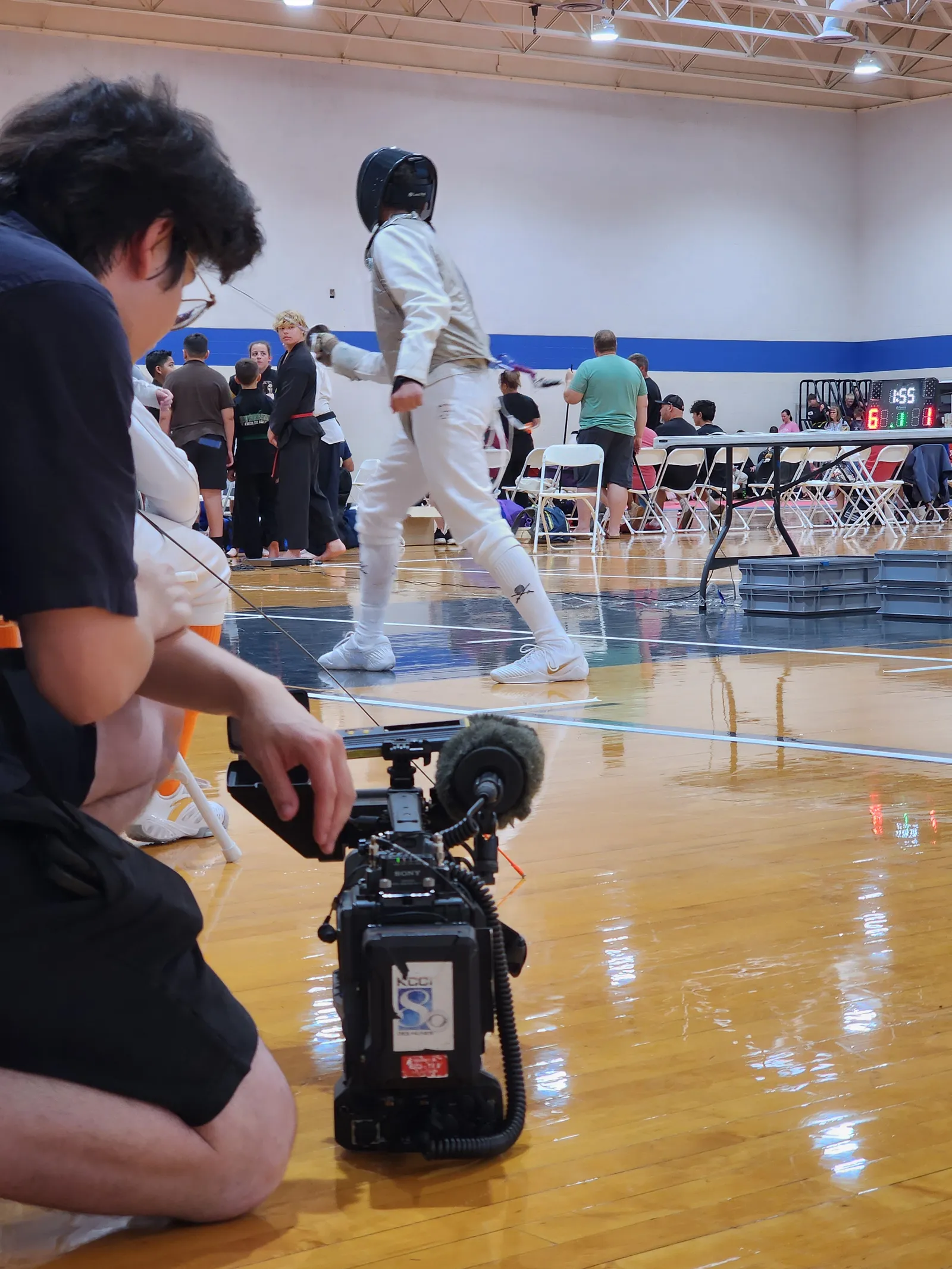 A KCCI Channel 8 camera operator kneels courtside filming a saber fencer walking back to the en-garde line.