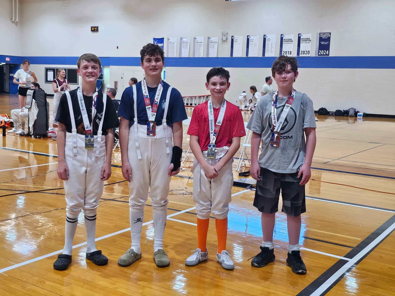 Four young foil fencers stand side by side on the gym floor wearing their Iowa Games medals.