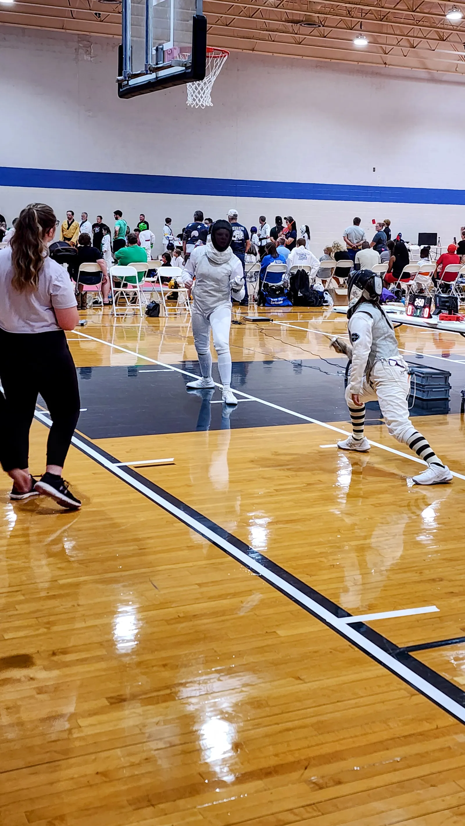 A fencer lunges toward an advancing opponent mid-bout while the referee watches from the near side of the strip.