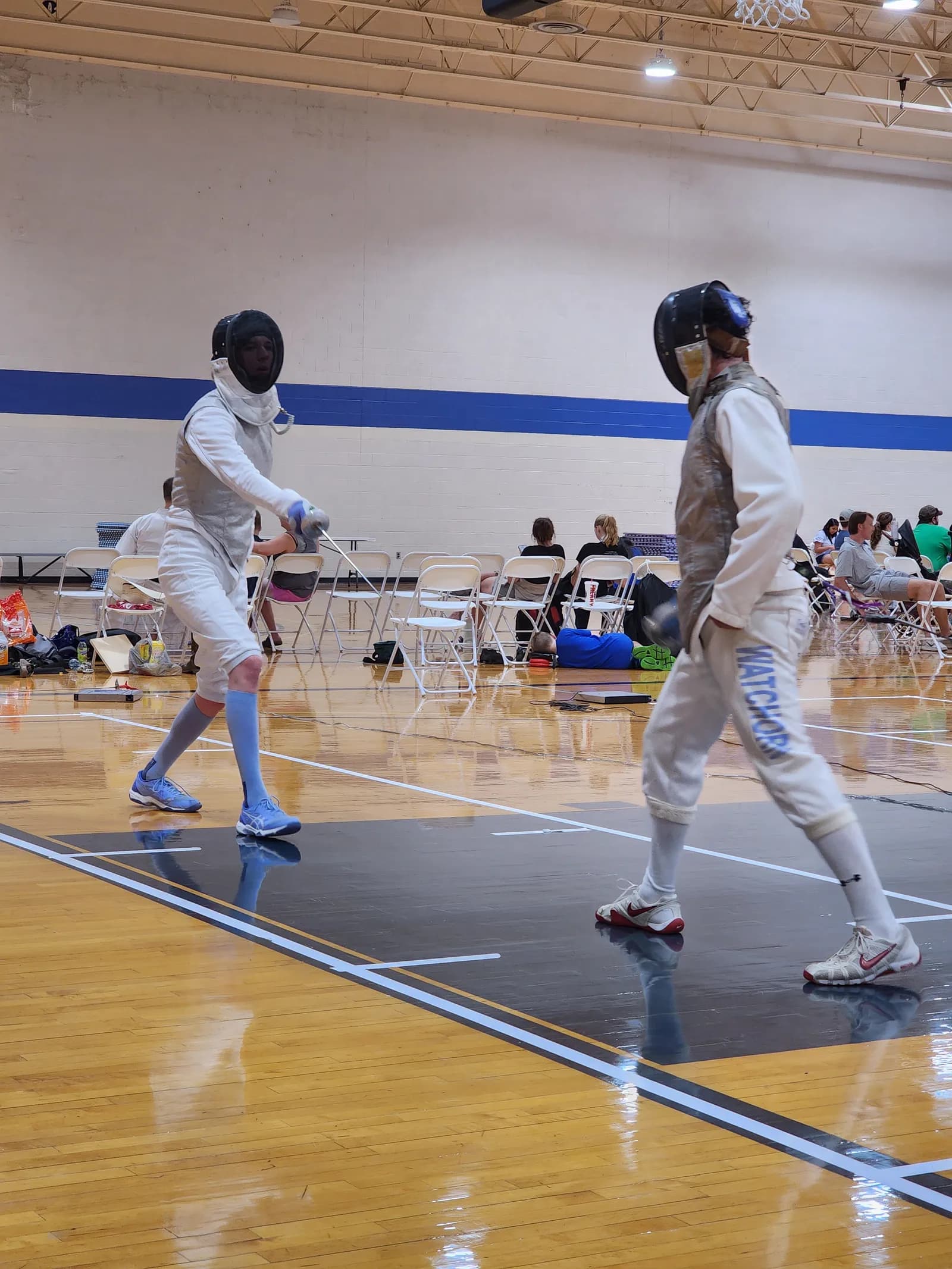 Two fencers in full kit square off at mid-strip, blades crossed, with spectators seated along the far wall.