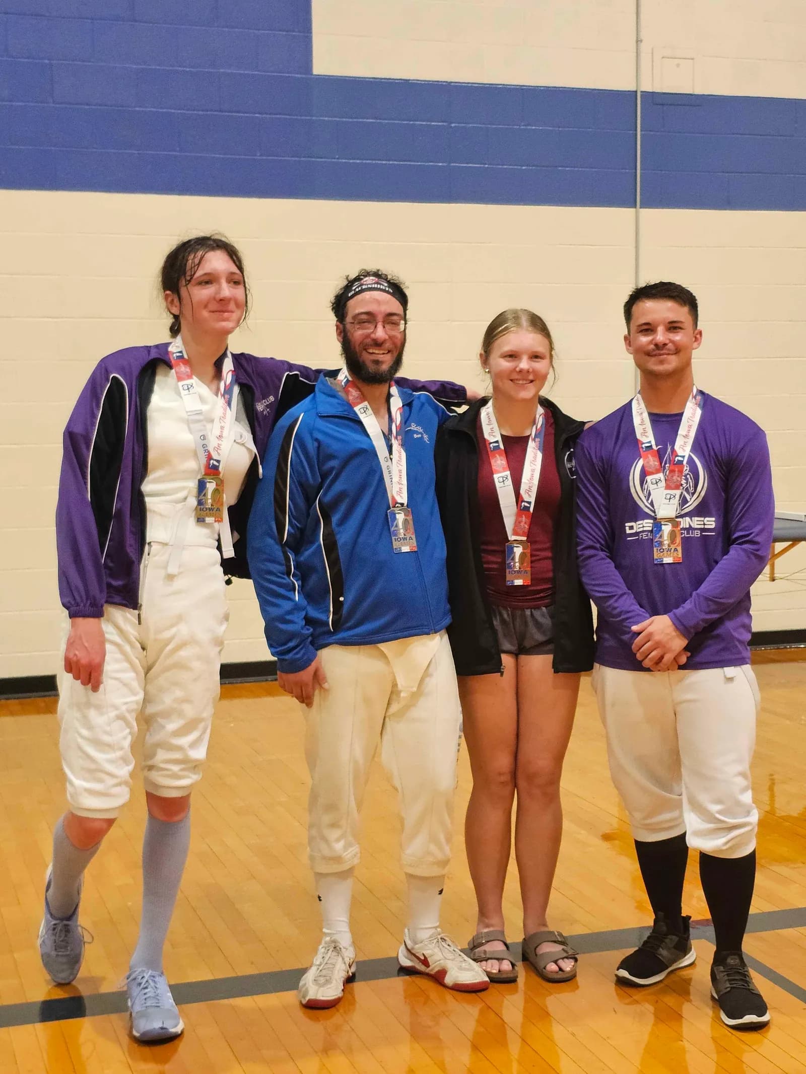 Four smiling fencers stand arm in arm wearing Iowa Games medals, one in a Des Moines Fencing Club shirt.